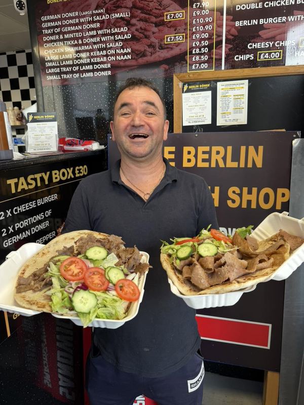 Berlin Doner staff holding two large doner kebabs on naan in front of the menu board